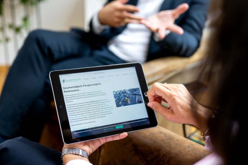 Eine Frau hält ein Tablet in der Hand auf der eine Website geöffnet ist. Ihr gegenüber sitzt ein Mann und gestikuliert mit den Händen.
