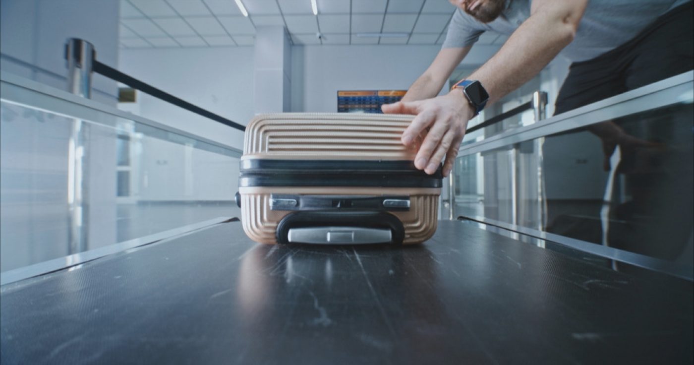Zu sehen ist die Fördertechnik: Die Hände eines Menschen im kurzärmeligen Hemd heben ein Gepäckstück von einem Kofferband. Ein Kontext zwischen Personalberatung und Fördertechnik.