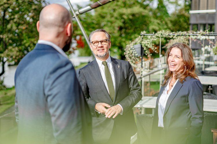 Geschäftsführer und Berater im Gespräch im Freien. Ein Business Meeting im Mittelstand. Drei Personen im Dialog, Eine Frau und ein Mann schauen einer dritten Person zu. Es scheint ein Businesskontext zu sein, die Stimmung ist gelöst und von hoher Aufmerksamkeit.