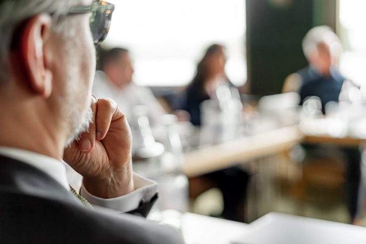 Ein Senior Manager im Anzug und mit Brille blickt im Rahmen eines Meetings in die Runde seiner Führungskräfte; macht den Eindruck des intensiven Denkens und hoher Konzentration